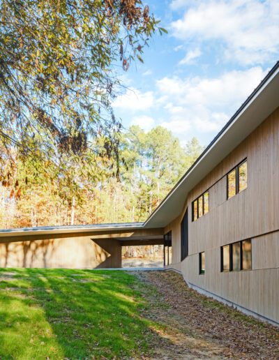 Modern wooden building with large windows in a wooded area under a blue sky. The surrounding landscape features green grass and trees with autumn foliage.