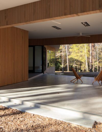 Two wooden chairs on a concrete patio beneath a modern wooden structure, overlooking a grassy area with trees in the background.