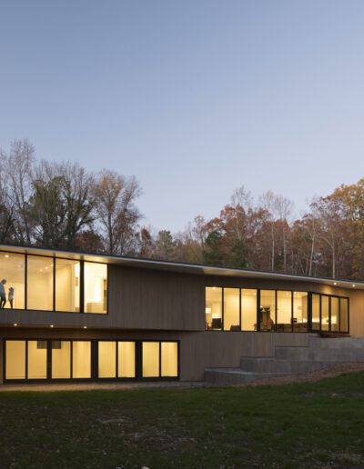 Modern, two-story house with large windows, lit from inside, surrounded by trees during dusk.