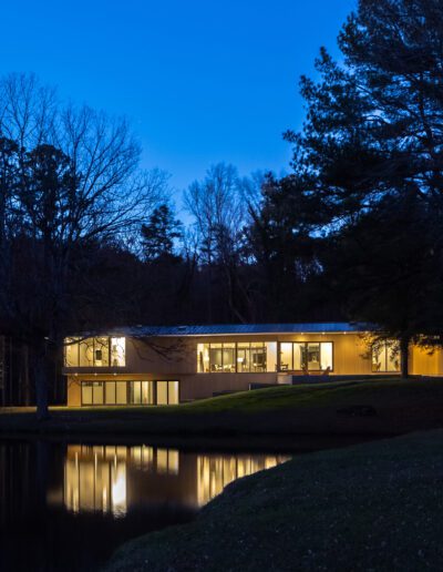 Modern house with large windows illuminated at dusk, surrounded by trees, reflecting in a calm pond in the foreground.