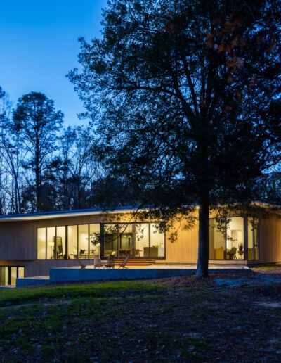 A modern, single-story house with large windows is illuminated at dusk, surrounded by trees and a grassy lawn.