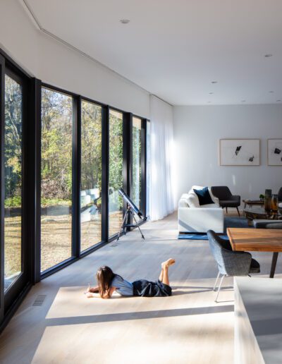 Child lying on the floor in a modern living room with large glass windows, wooden dining table, and white sofas; outdoor view of trees and lawn.