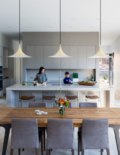 Modern kitchen and dining area with neutral tones. A person stands at a kitchen island, while two children are nearby. Large windows allow natural light to enter the space.
