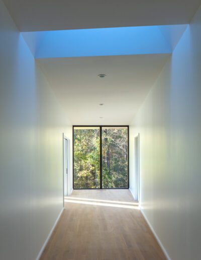 Minimalist hallway with a wooden floor, white walls, and a large window showing trees. Ceiling features skylights that allow natural light to illuminate the space.