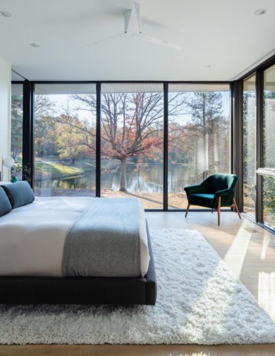 Modern bedroom with large windows overlooking a lake. Features a bed with gray and white bedding, a white rug, and a green chair. Bright natural light illuminates the room.