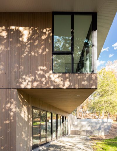 Modern building exterior with large windows, vertical wood paneling, and a cantilevered section. Sunlight filters through nearby trees, casting dappled shadows on the wall.