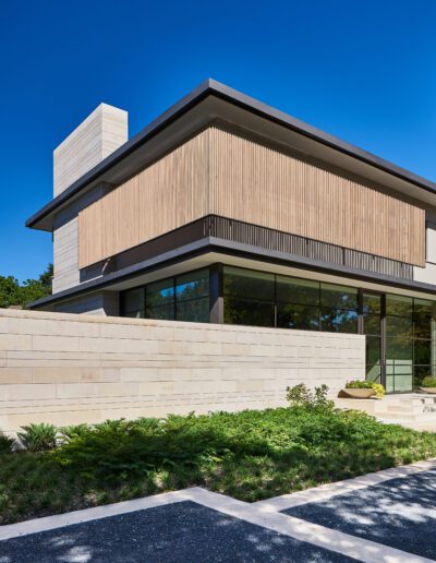Modern two-story building with large windows and wooden paneling. Stone exterior walls, a flat roof, and landscaped greenery in the foreground under a clear blue sky.