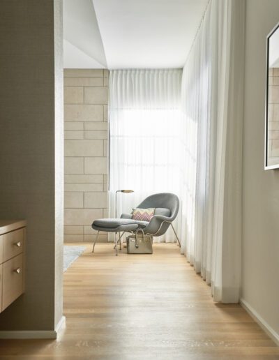 A minimalist room with a gray chair and footrest by a large window, a small round table with a lamp, and a bag on the floor. Light wood flooring and sheer curtains complete the setting.
