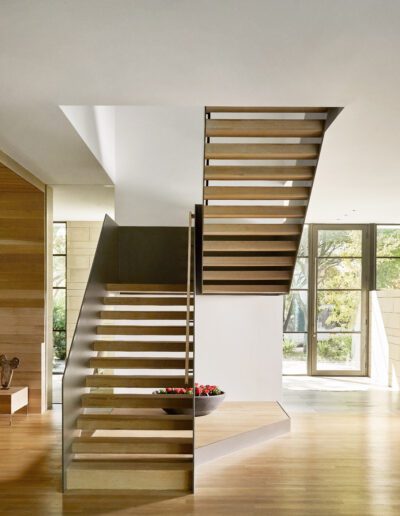 Modern interior features a wooden floating staircase with metal supports, leading to an upper floor. Large windows in the background provide natural light and a view of greenery.