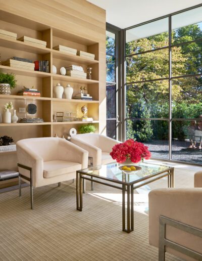 Modern living room with large windows, beige chairs, glass coffee table with pink flowers, and wooden shelves with books and decor. Sunlight streams through the windows, highlighting the garden.