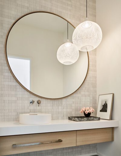 Modern bathroom with a round mirror, two pendant lights, a white sink on a wooden vanity, a small framed photo, and a vase with pink flowers.