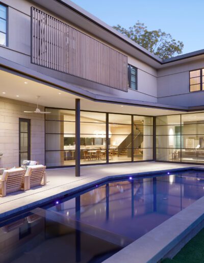 Modern house exterior with large windows, a covered patio with seating, and a rectangular swimming pool in the foreground.