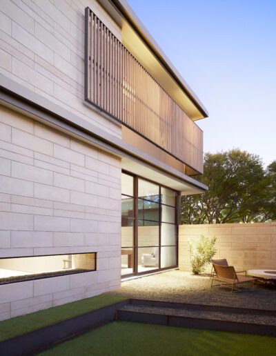 Modern house exterior with a minimalist design, featuring large glass windows, a wooden slatted screen, and a small outdoor seating area on gravel, bordered by a concrete fence.