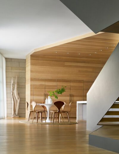 Modern dining area with a round table, six chairs, and a white vase with greenery. Wood-paneled walls, large sculpture, and minimal decor create a sleek and open space.