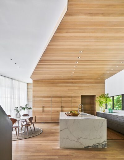 Modern kitchen with a marble island and wooden cabinetry. Large windows provide natural light. A dining table is visible in the open space, and plants add greenery to the decor.