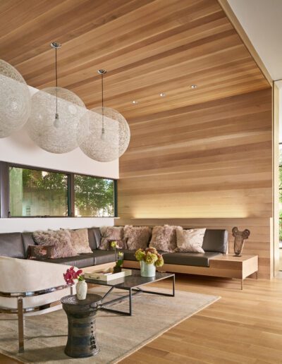 Modern living room with wooden walls and ceiling, a gray sectional sofa, round white pendant lights, a glass coffee table, and decorative vases. Large windows offer outdoor views.