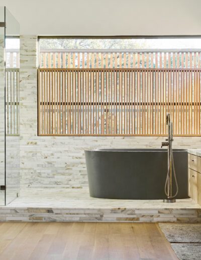 Modern bathroom with a freestanding black bathtub, glass-enclosed shower, wooden slatted window blinds, and dual sinks on a wooden vanity. Light wooden flooring throughout.