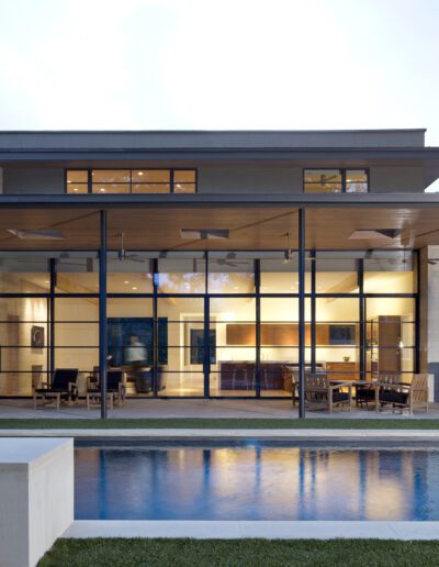 Modern house with large windows and a flat roof, featuring an outdoor pool in the foreground and visible interior spaces with furniture, surrounded by trees.