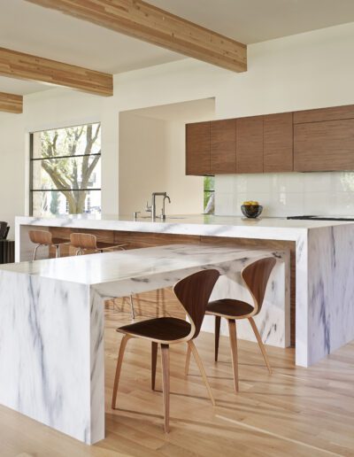 Modern kitchen with a marble island, wooden cabinets, and a dining area featuring brown chairs. Large windows provide natural light, and the room has wooden flooring and ceiling beams.