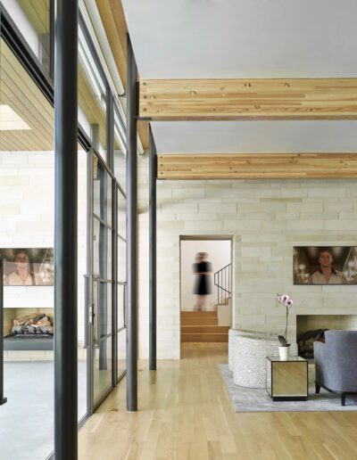 Modern living room with glass walls, wooden beams, and light stone accents. Features a gray sofa, fireplace, wall-mounted TV, and a visible stairway in the background.