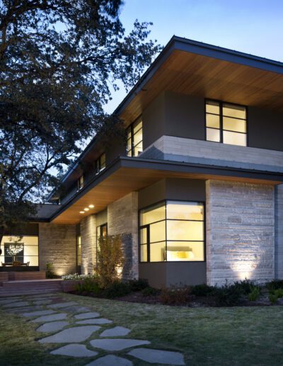Modern two-story house with large windows, a flat roof, and stone exterior. Illuminated entrance with a stone path and trees surrounding the property. Evening setting.