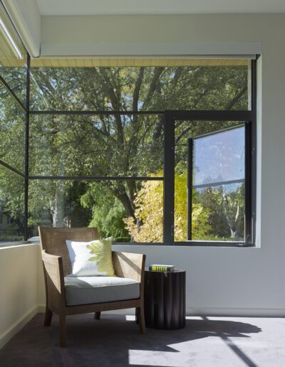 A cozy chair with a patterned cushion sits next to a small black side table by a window, offering a view of lush green trees outside.