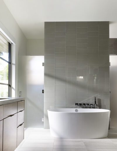 Modern bathroom with a white freestanding bathtub, gray-tiled wall, wooden vanity with drawers, and a large window providing natural light.