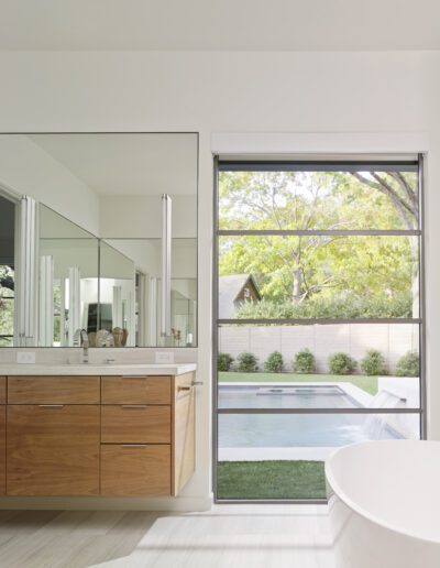 Modern bathroom with a large mirror above a wooden vanity, a freestanding bathtub, and a window offering a view of a pool and garden.