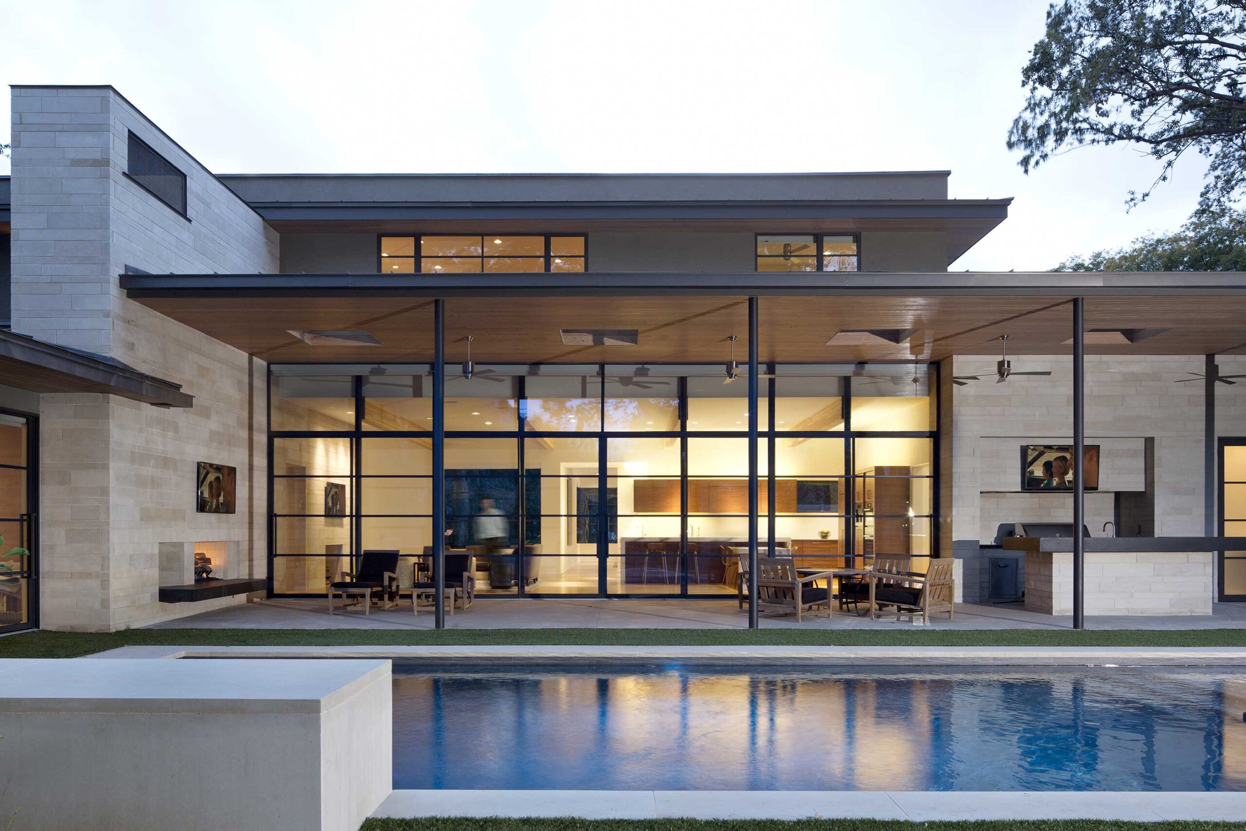 Modern house with large glass windows, displaying a well-lit interior. The backyard features a rectangular pool, patio with outdoor furniture, and a grill area under a covered patio.