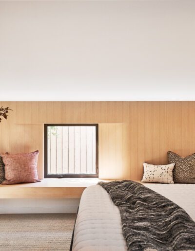 Modern bedroom with a wooden accent wall, window seat adorned with cushions, and a bed with patterned pillows and a textured throw. A vase with branches sits on a dresser.