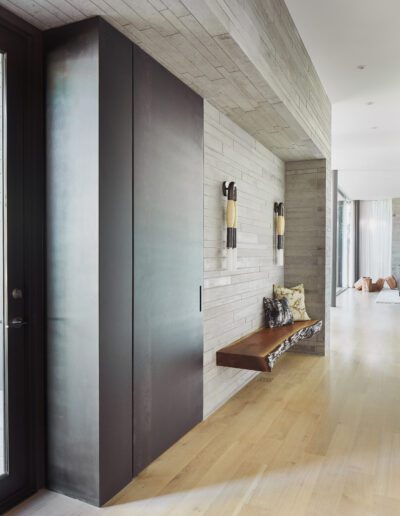 Modern hallway with light wood flooring, concrete walls, and a dark sliding door. A wooden bench features decorative pillows beneath two wall-mounted lights. Natural light enters from large windows.