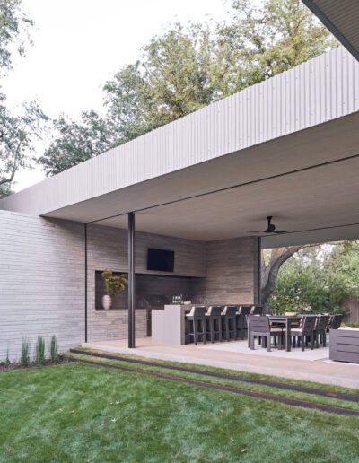 Outdoor covered patio with modern seating, dining tables, and a small bar area. Ceiling fans above, open view to a grassy yard and trees.
