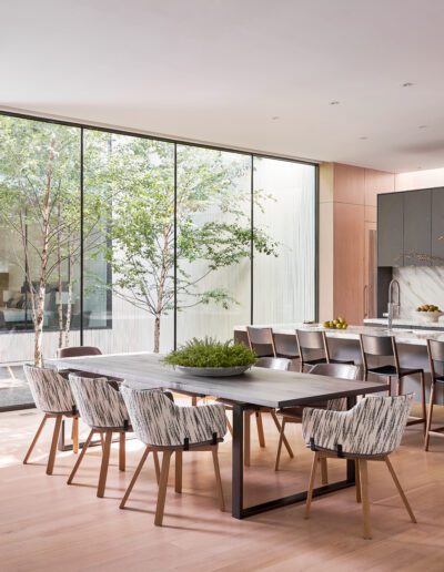 Modern kitchen with a large dining table and chairs. Glass wall reveals greenery outside. Kitchen island and sleek cabinets are visible under soft lighting.