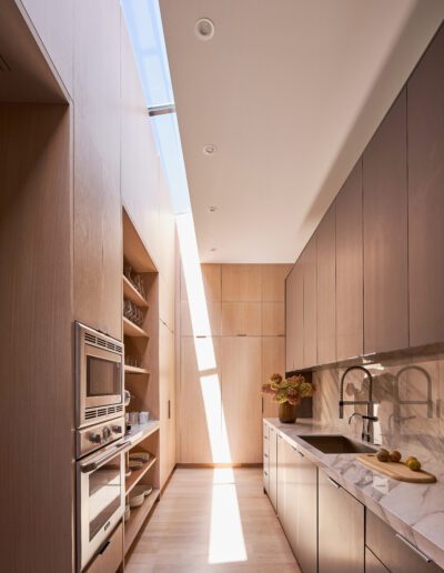Modern kitchen with light wood cabinets, marble countertops, built-in appliances, and a skylight. Natural light illuminates the space.