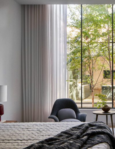 Bedroom with a large window, two blue chairs, a small table, a lamp, and a bed with a grey quilt. Draped curtains and a view of a tree outside.