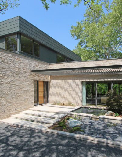 Modern house with stone and metal facade, large windows, and a flat roof, surrounded by trees. Gravel driveway and small landscaped garden in front.