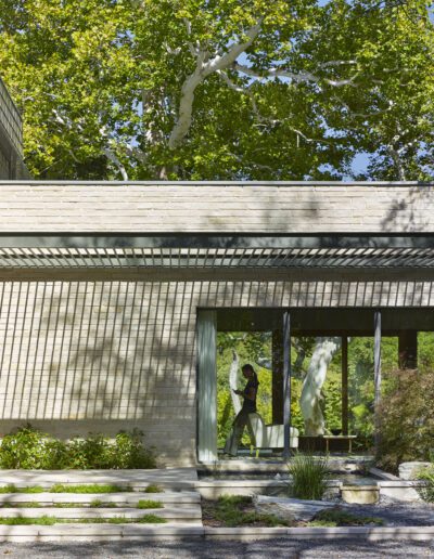 Modern house exterior with large windows, stone walls, and a pergola casting shadows. A person is visible inside, surrounded by trees and greenery in the background.