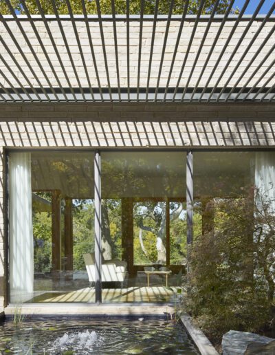Modern building facade with large glass windows reflecting trees. Overhead slatted structure casts striped shadows. Foreground features a pond and greenery.
