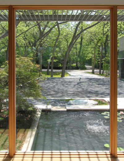 View through large window showing a tranquil garden with a pond, lily pads, and trees. Pathway and modern building visible in the background.