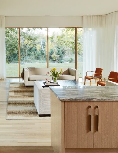 Modern living room with large windows showing a garden view, a beige sofa set, wooden chairs, a central table with flowers, and a kitchen island with a stone countertop in the foreground.