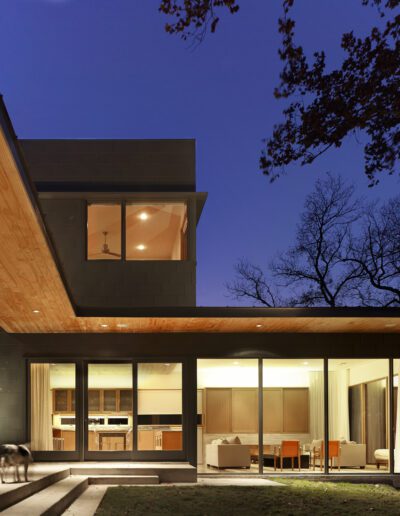 A modern house at dusk with large glass windows, revealing a lit interior. A person and a dog stand on the patio under a wooden overhang.