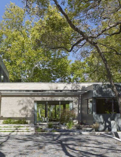 Modern house with large windows surrounded by trees, featuring a stone facade and gravel driveway under a clear blue sky.