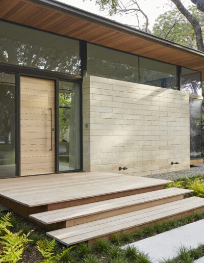 Modern house exterior with light stone walls and large glass windows. Wooden steps lead to a glass door, surrounded by green plants and trees.