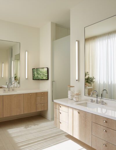 A modern bathroom with two wooden vanities, each featuring a mirror and vertical lights. A potted plant is on one vanity. Soft, natural light filters through a window with sheer curtains.