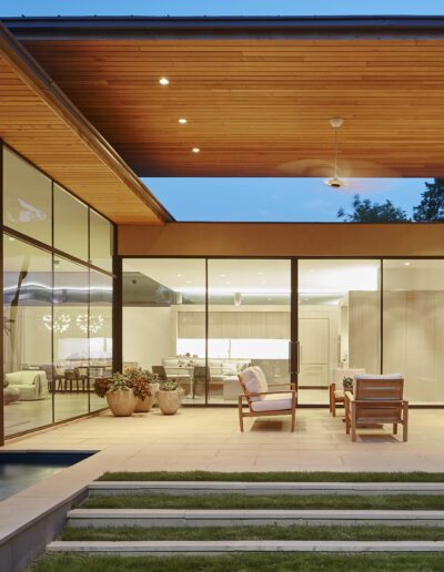 Modern house with large glass windows, patio with potted plants, wooden ceiling, and outdoor seating. Evening lighting creates a warm ambiance.