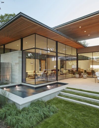 A modern house with large glass walls, a wooden ceiling, and a small reflecting pool surrounded by greenery. Outdoor seating is visible on the patio.