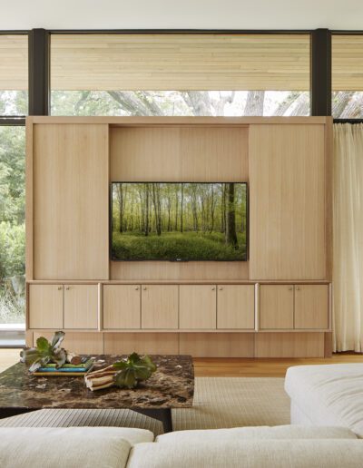 Modern living room with a large wood-paneled TV wall, beige sofas, armchairs, and a dark coffee table. Glass walls reveal a lush garden outside.