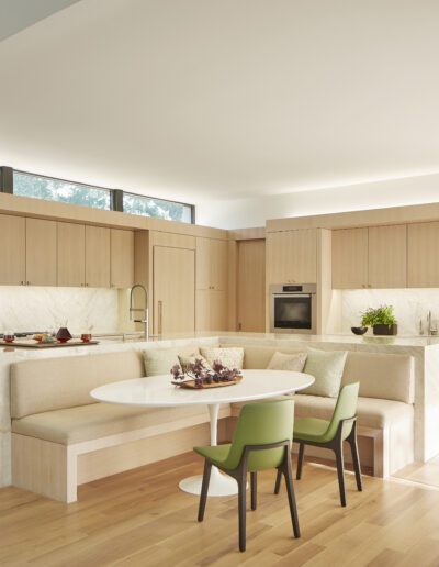 Modern kitchen with light wood cabinets and marble countertops. Built-in dining area with bench seating around a white table, accompanied by two green chairs. Bright, spacious, and minimalistic design.
