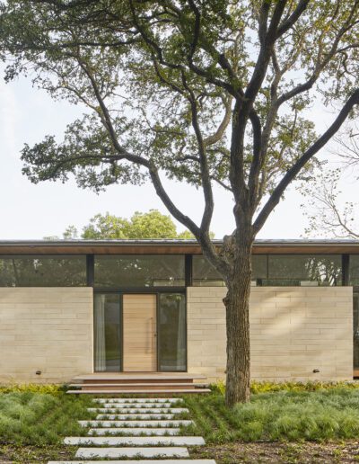 Modern one-story house with large glass windows and wooden accents, surrounded by trees and a landscaped garden, with a stone pathway leading to the entrance.