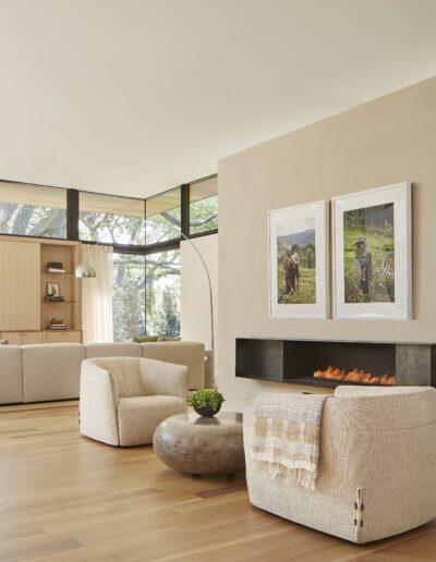 Spacious modern living room with light wood flooring, beige sofas, a central fireplace, and large windows. Two paintings hang above the fireplace. Natural light fills the room.